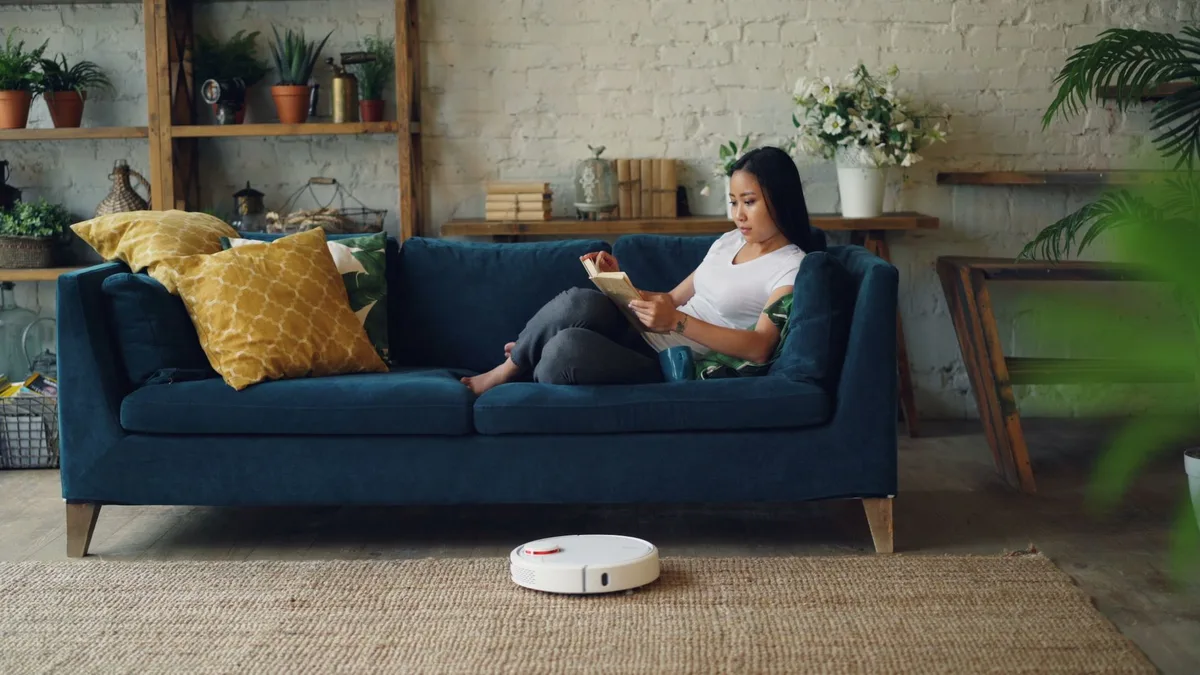Mujer leyendo en el sofá mientras una aspiradora robot limpia el living