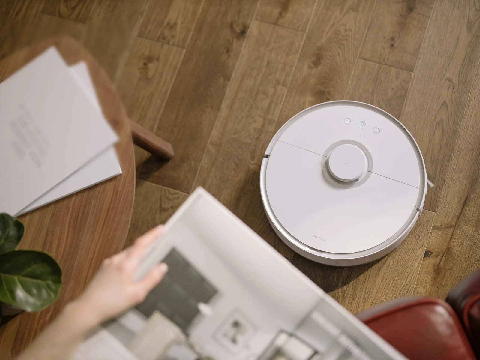 Aspiradora robot blanca limpiando el piso de madera de un living mientras una persona cuelga un cuadro en primer plano